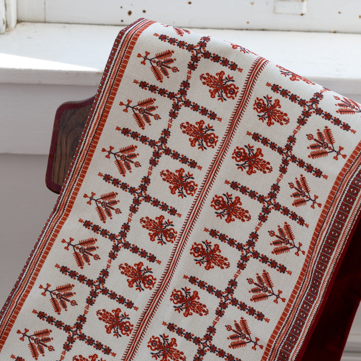 Decorative red and white Palestinian tatreez patterned table runner draped over a chair by a window.